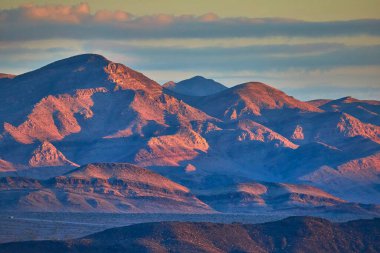 Hindistan Pınarları 'nın yakınındaki engebeli Nevada dağlarının üzerinde ılık altın saat ışığı süzülüyor. Yumuşak bulutlar yükseliyor. Gün batımı kurak arazide dramatik gölgeler yaratır ve doğayı güçlendirir.