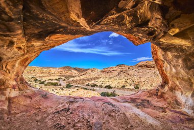 Çamurlu Dağ 'daki kayalık bir mağaradan baktığımızda parlak mavi gökyüzünün altındaki güneş ışığıyla aydınlanmış Moapa Vadisi manzarasını görüyoruz. Bu çarpıcı tepede dokulu kaya duvarlar kurak Nevada arazisini ve uzak tepeleri çerçeveliyor.