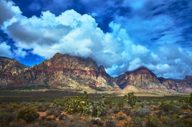 Las Vegas Nevada yakınlarındaki Red Rock Kanyonu 'nun kırmızı kumtaşı dağları üzerinde çarpıcı bulutlar yuvarlanıyor. Geniş açıda parlak bir gökyüzü altında canlı kayalık kayalıklar ve vahşi Mojave çöl bitkileri sergileniyor..