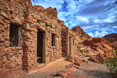 Kulübe Nevada Moapa Vadisi yakınlarındaki Ateş Vadisi 'nde dramatik kızıl kaya oluşumları arasında yer alıyor. Güneş ışığı, çarpıcı mavi gökyüzünün altındaki engebeli taş duvarları ve canlı çöl manzarasını vurgular..