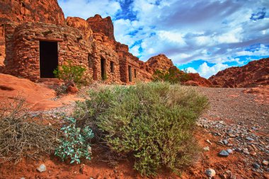 Ateş Vadisi 'nde kırmızı kumtaşı ve çöl bitkileriyle çevrili tarihi bir taş kulübe duruyor. Dramatik bulutlar ve uzak Moapa Vadisi ayarı bir macera ve tarih duygusu yaratır.