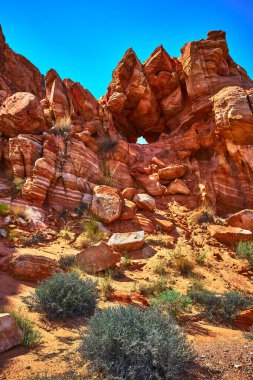 Sunlit red sandstone and a dramatic natural arch rise above scattered boulders and desert plants in Nevadas Moapa Valley. The rugged landscape along the Bitter Springs Trail captures the wild spirit