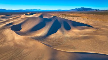 Nevada 'daki Büyük Kumul' un havadan görünüşü dramatik kum tepesi şekilleri ve açık mavi gökyüzünün altındaki çöl kumları. Lastik izleri ve uzak dağlar olay yerine derinlik ve macera ekler..
