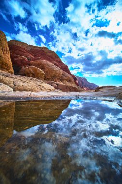 Las Vegas Nevada yakınlarındaki Red Rock Kanyonu 'ndaki sakin bir su birikintisinin üzerinde Calico' nun canlı kumtaşı dağları yükseliyor. Gökyüzü ve kaya oluşumları güzel bir şekilde suya yansıyarak dramatik bir çöl oluşturur.