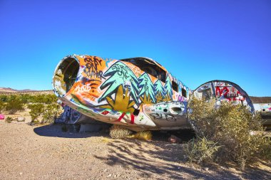Canlı, terk edilmiş bir uçak, Beatty yakınlarındaki Nevada çölünde renkli graffitilerle kaplıdır. Güneş ışığı bulutsuz bir Birleşik Devletler gökyüzünün altında uçak enkazı ve çöl fırçasını vurguluyor..