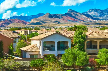 Las Vegas 'ın panoramik manzarasında arka planda dramatik Kızıl Kaya dağlarının yükseldiği yemyeşil alanlar arasında yuva yapmış klasik evler sergileniyor. Parlak gökyüzü ve canlı renkler güçlendirir