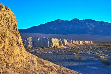 Sıcak güneş ışığı, Las Vegas Nevada yakınlarındaki Tule Springs Fosil Yatağı 'ndaki fosil yatak oluşumlarını ve çöl bitkilerini vurguluyor. Uzak dağlar ve berrak mavi gökyüzü dramatik bir altın saat yaratır.