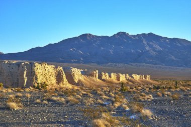 Sıcak güneş ışığı, Nevada 'daki Tule Springs Fosil Yatağı' nın kayalık arazisini ve aşınmış kayalıkları aydınlatır. Uzak dağlar, Las Vegas yakınlarındaki açık mavi gökyüzünün altında dramatik gölgeler bırakır.