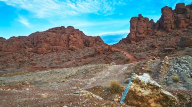 Las Vegas Nevada yakınlarındaki bir çöl arazisinden yükselen engebeli kırmızı kaya oluşumları Lava Butte 'a doğru dramatik bir iz bırakıyor. Sahne gün ışığıyla aydınlanır ve canlı mavi bir gökyüzü tarafından çerçevelenir..