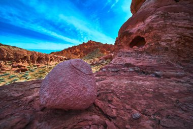 Las Vegas Nevada yakınlarındaki Ateş Kasesi 'nde parlak mavi gökyüzünün altında kırmızı kumtaşı oluşumlarının ortasında çarpıcı bir kaya parçası duruyor. Dramatik çöl manzarası, doğanın güzelliğini vurgular.