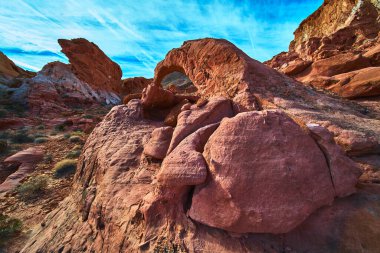 Güneş ışığı Las Vegas yakınlarındaki Ateş Kasesi 'ndeki etkileyici kumtaşı kemeri ve koyu kırmızı kaya oluşumlarını vurguluyor. Canlı mavi gökyüzü ve çöl bitkisi çarpıcı bir doğal manzara yaratıyor..