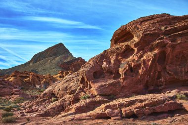 Canlı kırmızı kaya oluşumları arka planda engebeli bir dağ zirvesi ve ilham verici bir çöl sahnesi yaratan mavi gökyüzü ile Las Vegas Nevada yakınlarındaki Ateş Kasesi 'ne hakim..