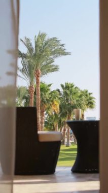 dikey video. Ön planda, rüzgardan sarkan perdeler, masada bir rattan sandalye ve bir masa, beyaz bir fincan ve saydam boş bir kül tablası, odak dışı. Avuç içi ve mavi gökyüzünün arka planında. Günaydın, Mısır, tam yol, ses yok.