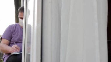 Tıbbi maskeli bir adam pencere kenarına oturur ve bir defterde kalemle yazar, sonra defterden bir çarşaf yırtıp kenara atar. kamera hareketi sağdan sola.