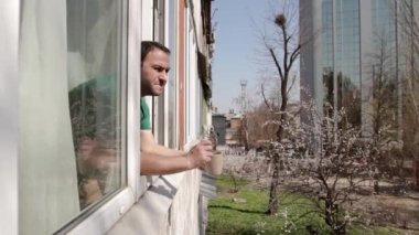 Bir adam açık pencerede sol elinde bej bir bardakla beliriyor, sağ eliyle selam veriyor ve geri dönüyor. Kameranın sağdan sola doğru hareket etmesi