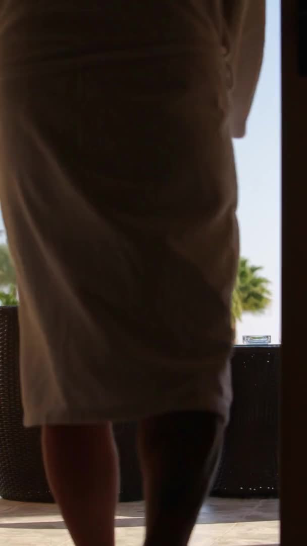 一个身穿白色长袍 头戴白色杯子的男人离开了阳台上的房间 坐在扶手椅上 喝着杯子里的水 窗帘在前边摇曳 背景中的棕榈树 垂直的视频 — 图库视频影像