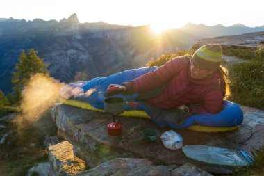 A woman cooking breakfast from her bivouac on a beautiful morning in the mountains. clipart