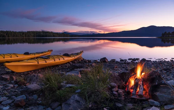 Alacakaranlıkta iki kanoyla gölün kıyısında kamp ateşi. İsveç.