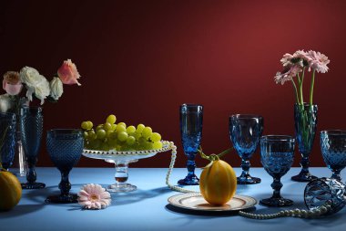 Beyaz tabaklar ve mumlarla süslenmiş mavi bardaklar, yeşil yapraklar, çiçek, meyveli katlı tepsi..