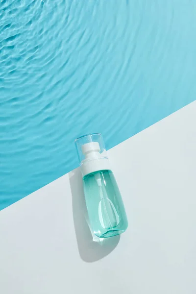 Blue cosmetic bottle on white square podium and the blue water surface. Blank label with texture for branding mock-up. Flat lay, top view.