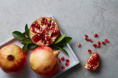 Olgun nar meyvesi ve yarım nar tohumu, gri beton arka planda yapraklı nar tohumu. Beyaz tabakta taze nar var. Vegan, sağlıklı beslenme konsepti. Üst görünüm.