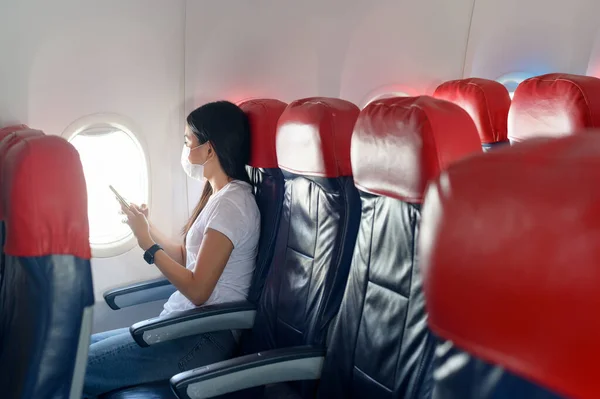A traveling woman wearing protective mask onboard in the aircraft using smartphone, travel under Covid-19 pandemic, safety travels, social distancing protocol, New normal travel concept