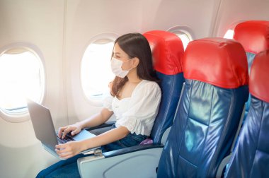 Yüz maskesi takan genç bir kadın gemide dizüstü bilgisayar kullanıyor. Covid-19 salgınından sonra normal bir seyahat.