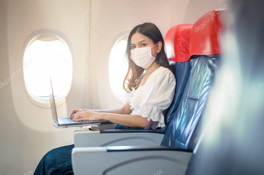 A young woman wearing face mask is using laptop onboard, New normal travel after covid-19 pandemic concept	