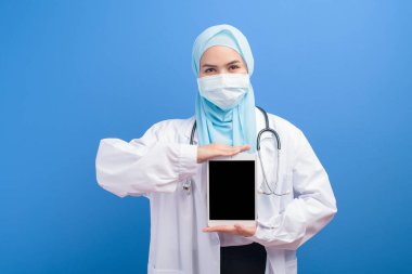 Türbanlı kadın bir müslüman doktor mavi arka plan stüdyosunun üzerinde tablet kullanarak cerrahi maske takıyor..