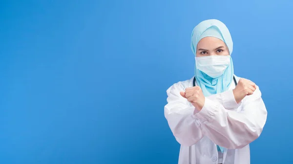 A female muslim doctor with hijab wearing a surgical mask over blue background studio.