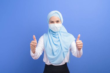 Mavi arka plan stüdyosu üzerinde cerrahi maske takan tesettürlü genç Müslüman bir kadın..