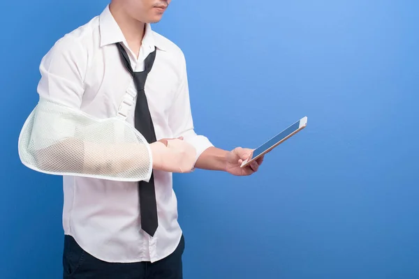 A young businessman with an injured arm in a sling using a tablet over blue background in studio, insurance and healthcare concept