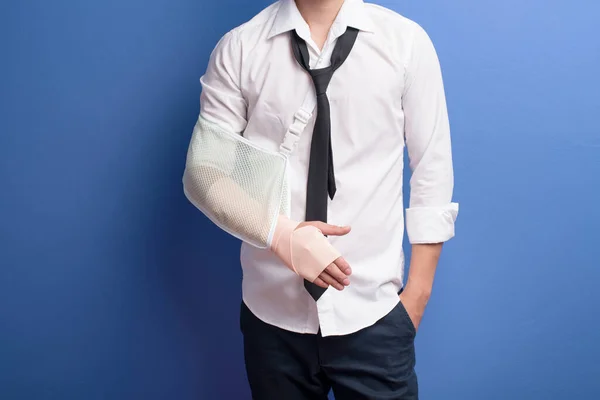 A young businessman with an injured arm in a sling over blue background in studio, insurance and healthcare concept