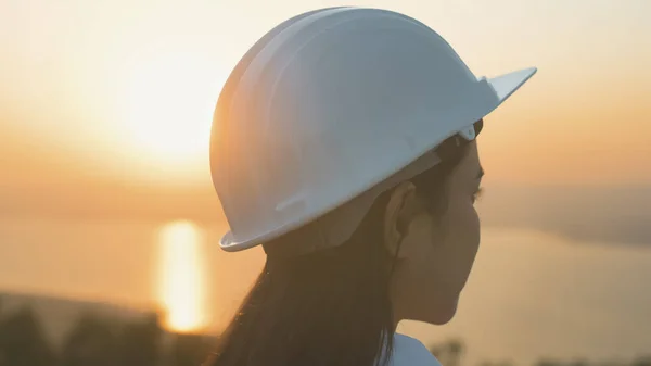 a woman engineer is putting a protective helmet on her head at sunset.