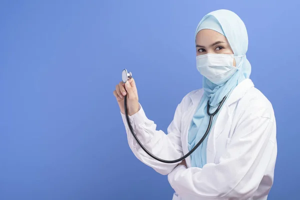 A female muslim doctor with hijab wearing a surgical mask over blue background studio.