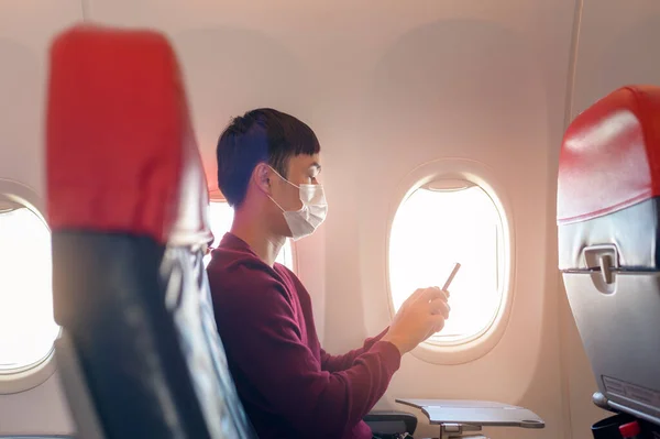 A traveling man wearing protective mask onboard in the aircraft using smartphone, travel under Covid-19 pandemic, safety travels, social distancing protocol, New normal travel concept