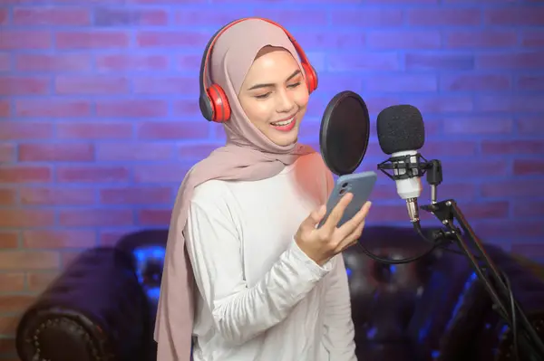 一名年轻 面带微笑的穆斯林女歌手 戴着耳机 带着话筒 在一个灯光艳丽的音乐工作室里录制歌曲 — 图库照片
