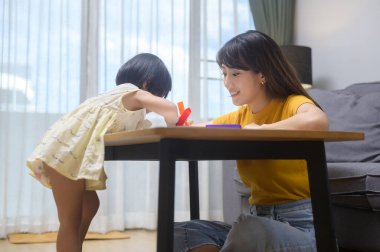 Genç bir anne, evdeki oturma odasında renkli kalemlerle resim çizmesine yardım ediyor..