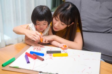 Genç bir anne, evdeki oturma odasında renkli kalemlerle resim çizmesine yardım ediyor..