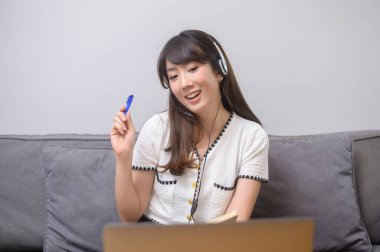 Kulaklık takan genç ve güzel bir kadın evdeki bilgisayarla video konferans görüşmesi yapıyor. 