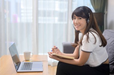 Kulaklık takan genç ve güzel bir kadın evdeki bilgisayarla video konferans görüşmesi yapıyor. 