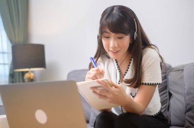 Kulaklık takan genç ve güzel bir kadın evdeki bilgisayarla video konferans görüşmesi yapıyor. 