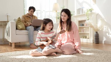 Anne ve babası oturma odasında keyfine bakarken kız ukulele çalıyor..