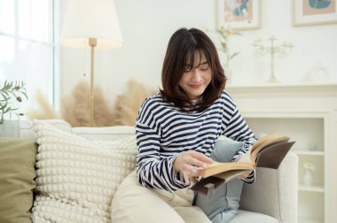 Genç bir kadın kanepede oturuyor, rahat bir ev ortamında bir kitaba dalmış..