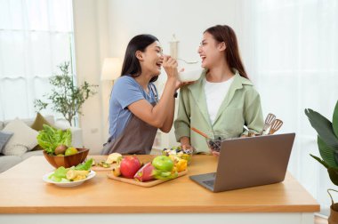 İki arkadaş gülümsüyor ve güneşli bir mutfakta yemek fikirleri için dizüstü bilgisayara bakıyor..