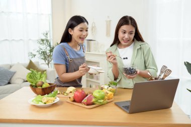 İki arkadaş gülümsüyor ve güneşli bir mutfakta yemek fikirleri için dizüstü bilgisayara bakıyor..