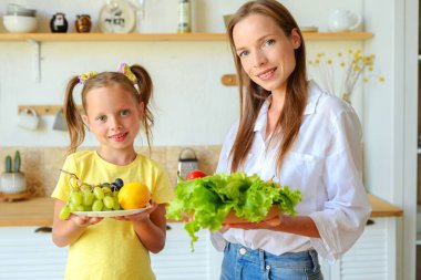 Anne ve küçük kızı mutfakta sağlıklı yiyecekler hazırlayıp çay içiyorlar.