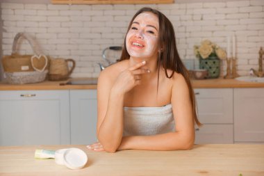 Çekici genç bir kadın yüz kremi sürüyor. Havluya sarılı güzel, sağlıklı bir kadın. Nemlendirici krem sürüyor.