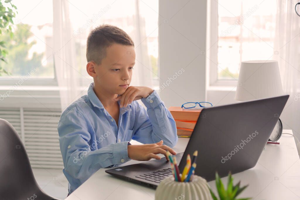 Schoolboy studies at home while sitting at table in front of laptop and listens attentively to information
