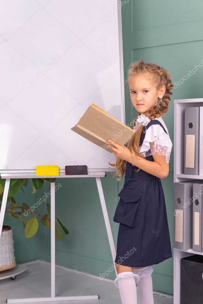 Back to school and happy time! A cute hardworking child is sitting at a table indoors. girl studying in the classroom on the background of the blackboard. The girl is reading a book.